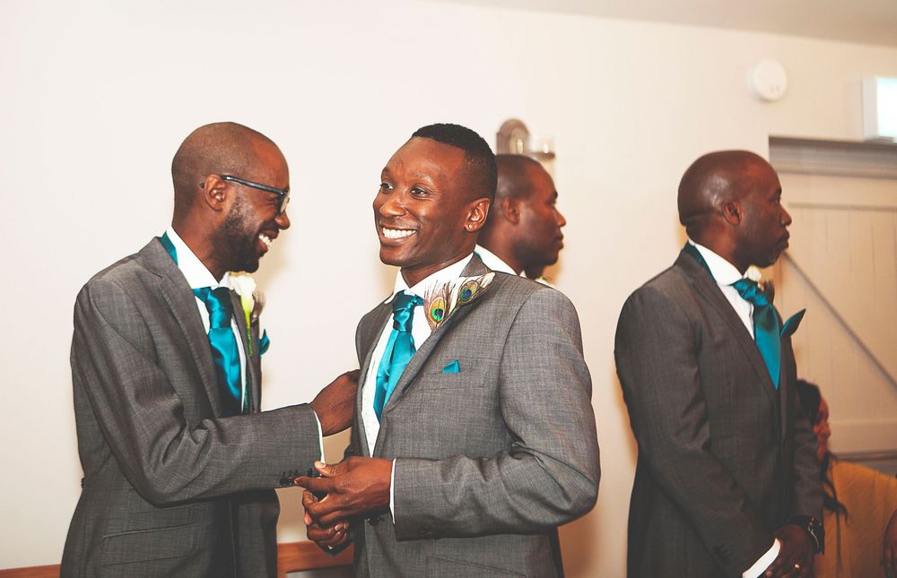 Groomsmen in grey suits with teal ties laughing and shaking hands at a wedding celebration. At Oaks farm wedding venue in Surrey