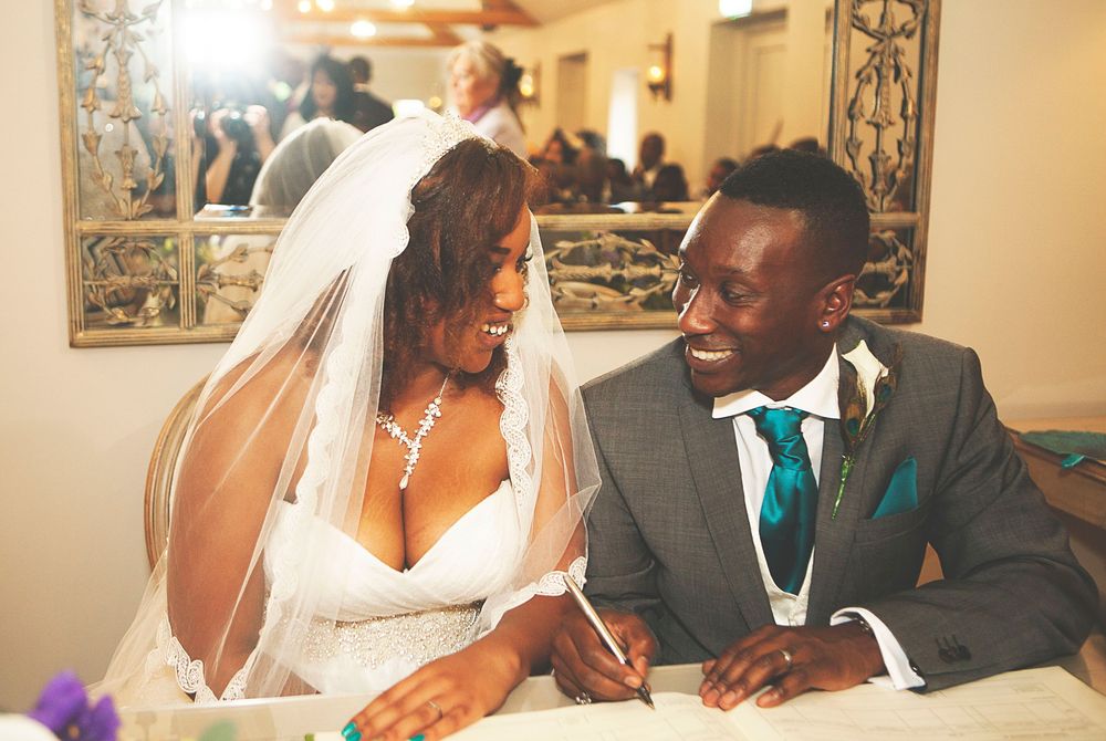 Smiling bride and groom in wedding attire signing marriage documents at a table during their ceremony. At Oaks farm wedding venue in Surrey.