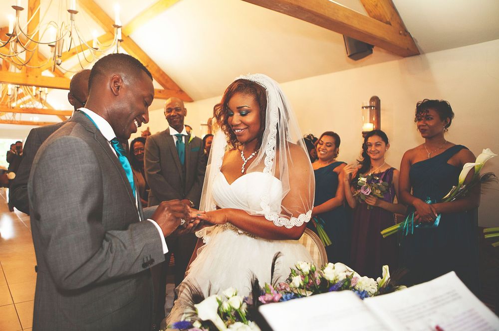 Bride and groom exchange rings during wedding ceremony with bridesmaids in teal dresses in background. At Oaks farm wedding venue in Surrey.