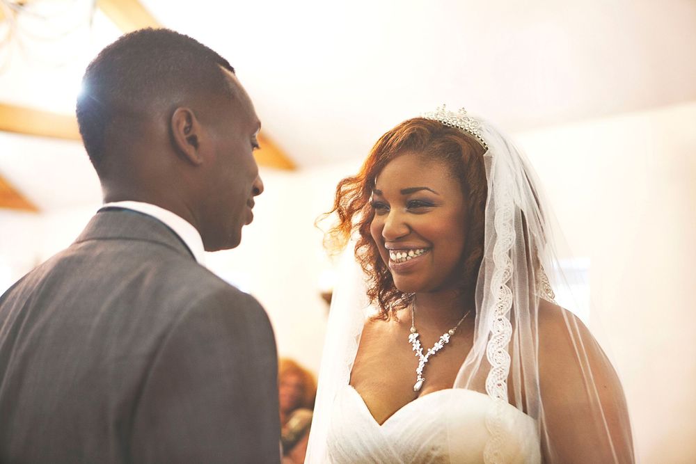 Smiling bride in white gown and veil exchanges vows with groom in grey suit during wedding ceremony. At Oaks farm wedding venue in Surrey