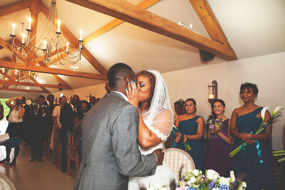 A bride and groom share their first kiss at a wedding ceremony in a rustic venue with wooden beams and chandelier at Oaks farm.