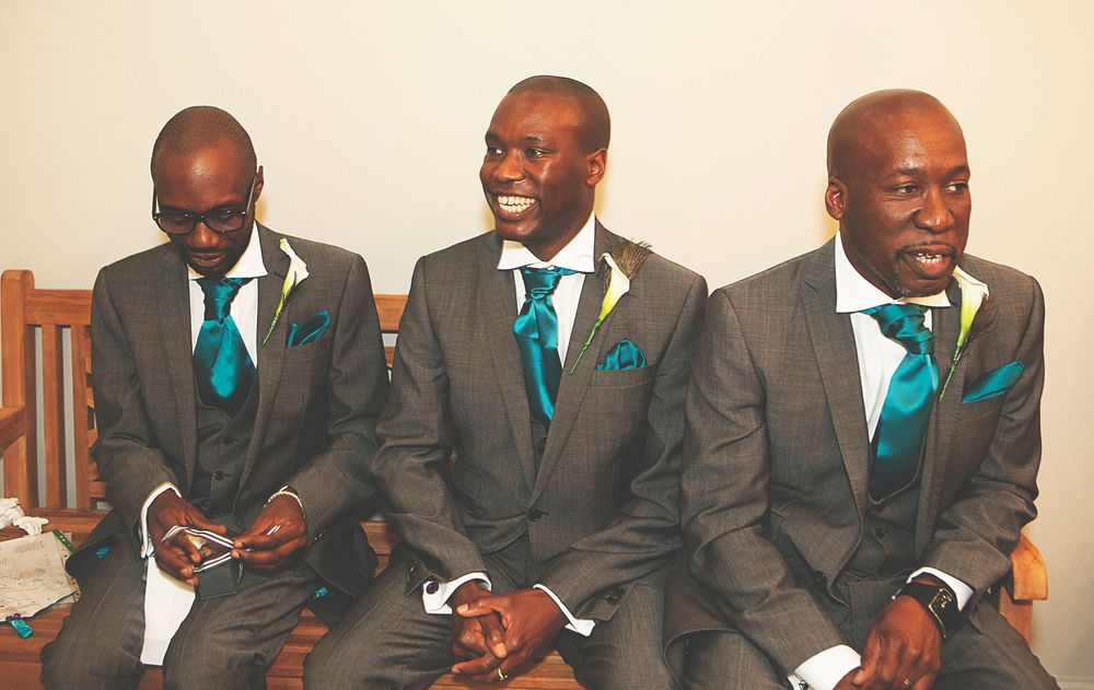 Three groomsmen in gray suits with teal ties and pocket squares seated together smiling at a wedding ceremony. At Oaks farm wedding venue.