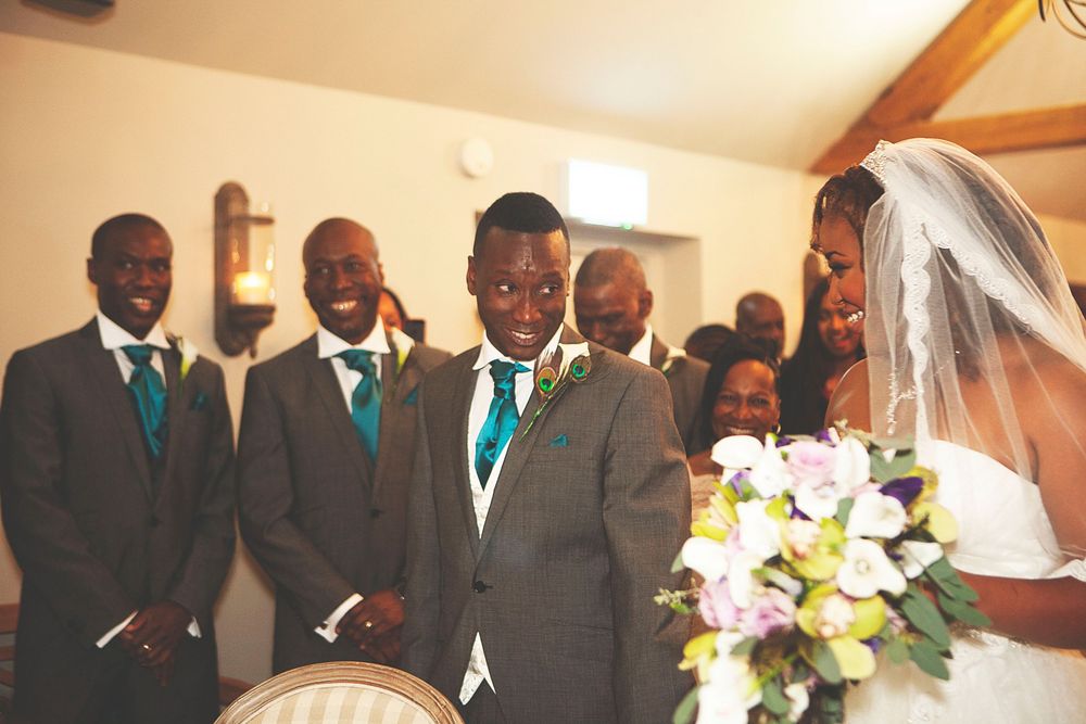 Groom and groomsmen in grey suits with teal ties stand at wedding altar as bride in white veil approaches. At Oaks farm wedding venue.