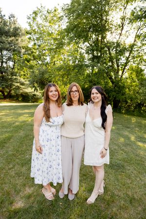 Three smiling women posing together outdoors on green grass with lush trees in the background.