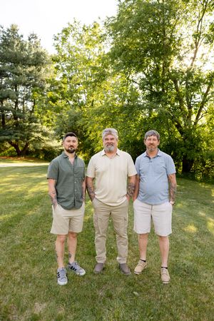 Three generations of men stand together smiling on green lawn with trees in golden sunlight.
