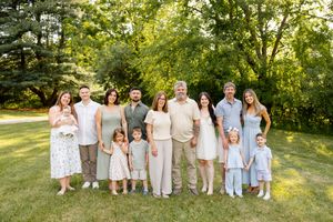 Large family group portrait outdoors on green lawn with tall trees, featuring three generations in coordinated neutral outfits.