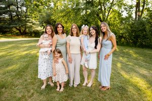 Group of seven women and children posing outdoors on green grass, wearing pastel outfits, smiling together on a sunny day.