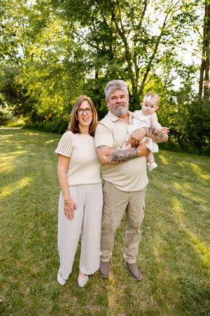 Smiling grandparents holding a baby outdoors on green grass with lush trees in the background on a sunny day.