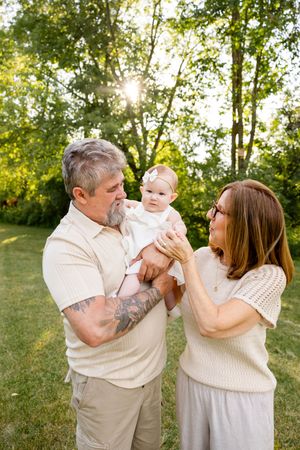 Grandparents with tattoos lovingly hold smiling baby girl in white dress in sunlit green park.