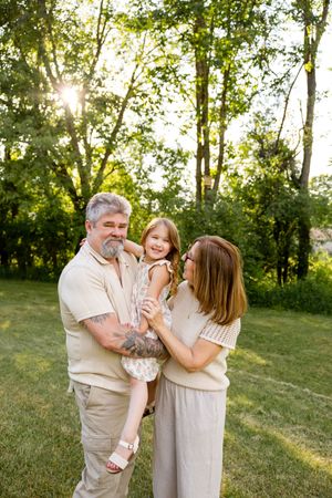 A happy family with a young girl poses outdoors in golden sunlight surrounded by lush green trees.