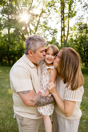 Grandparents kissing happy toddler girl on cheeks outdoors with sunlight streaming through trees.