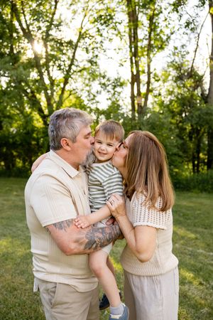 Grandparents both kiss cheeks of happy toddler boy in striped shirt in sunny green park.