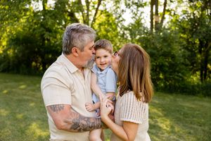 A couple kisses their smiling toddler boy outdoors in a lush green park setting.
