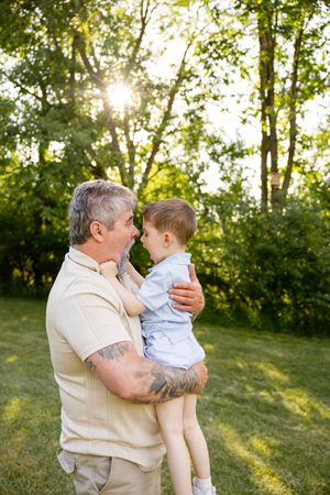 A tattooed grandfather holds his young grandson in a sunny green park, sharing a tender nose-to-nose moment.