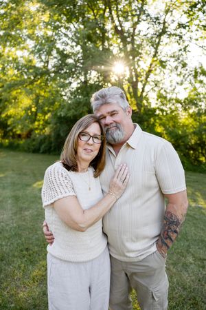 Affectionate middle-aged couple embracing outdoors with sunlight filtering through green trees behind them.