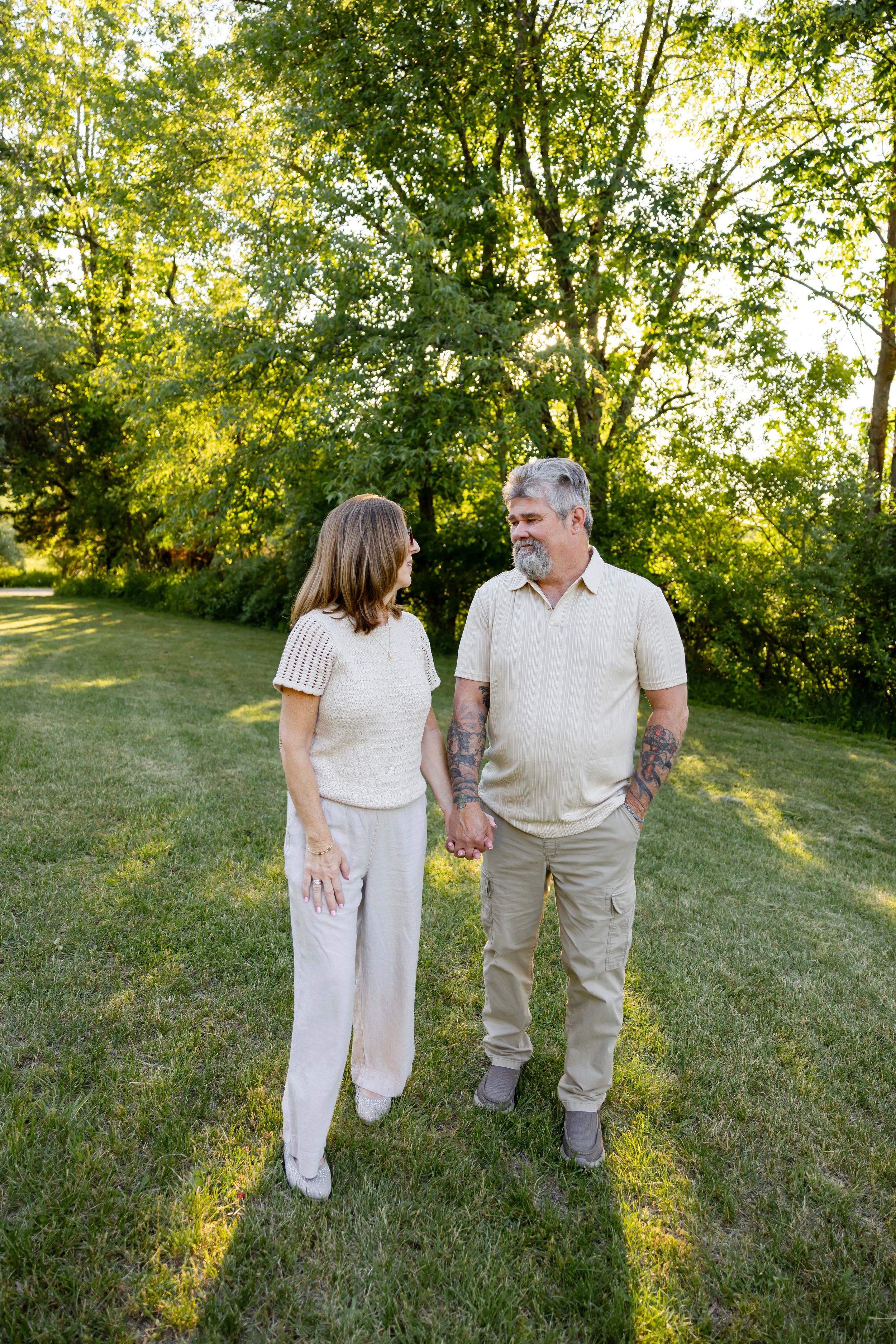 A middle-aged couple holds hands and gazes at each other while walking on a sunlit green lawn.