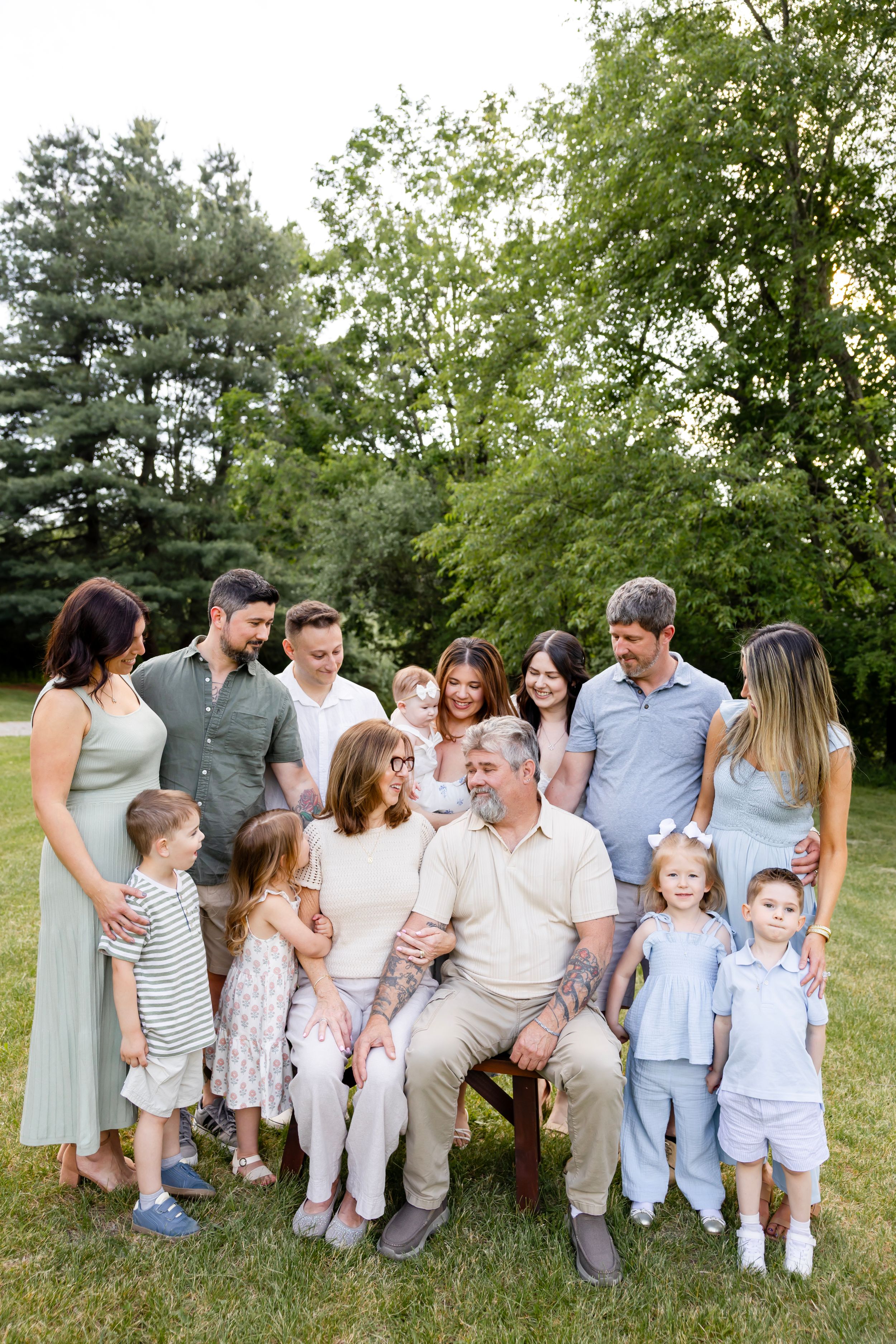 Large multigenerational family gathering outdoors on green lawn with tall trees in background during golden hour. NJ family photographer