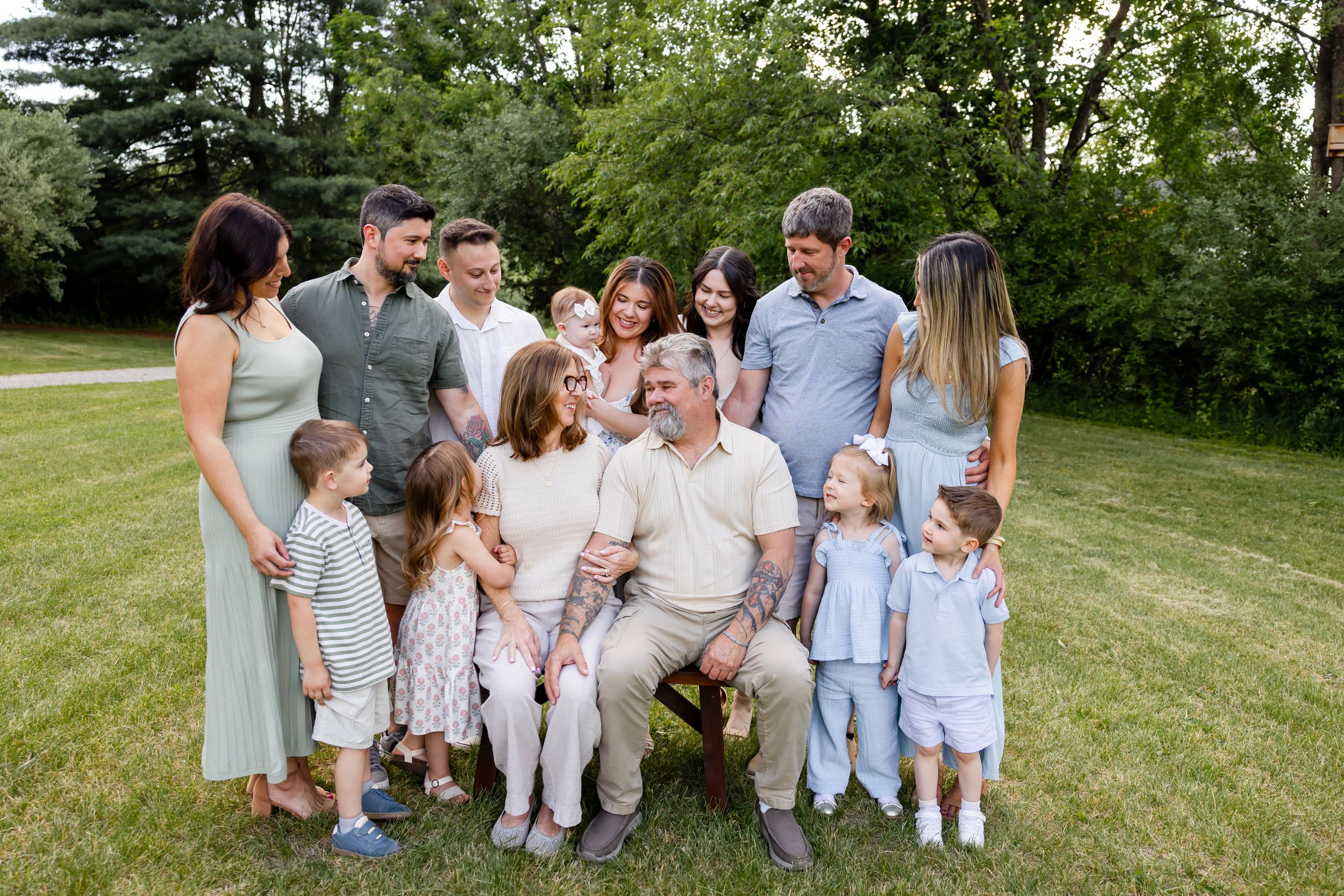 Large multigenerational family posing together outdoors on green grass surrounded by lush trees during a family portrait session.