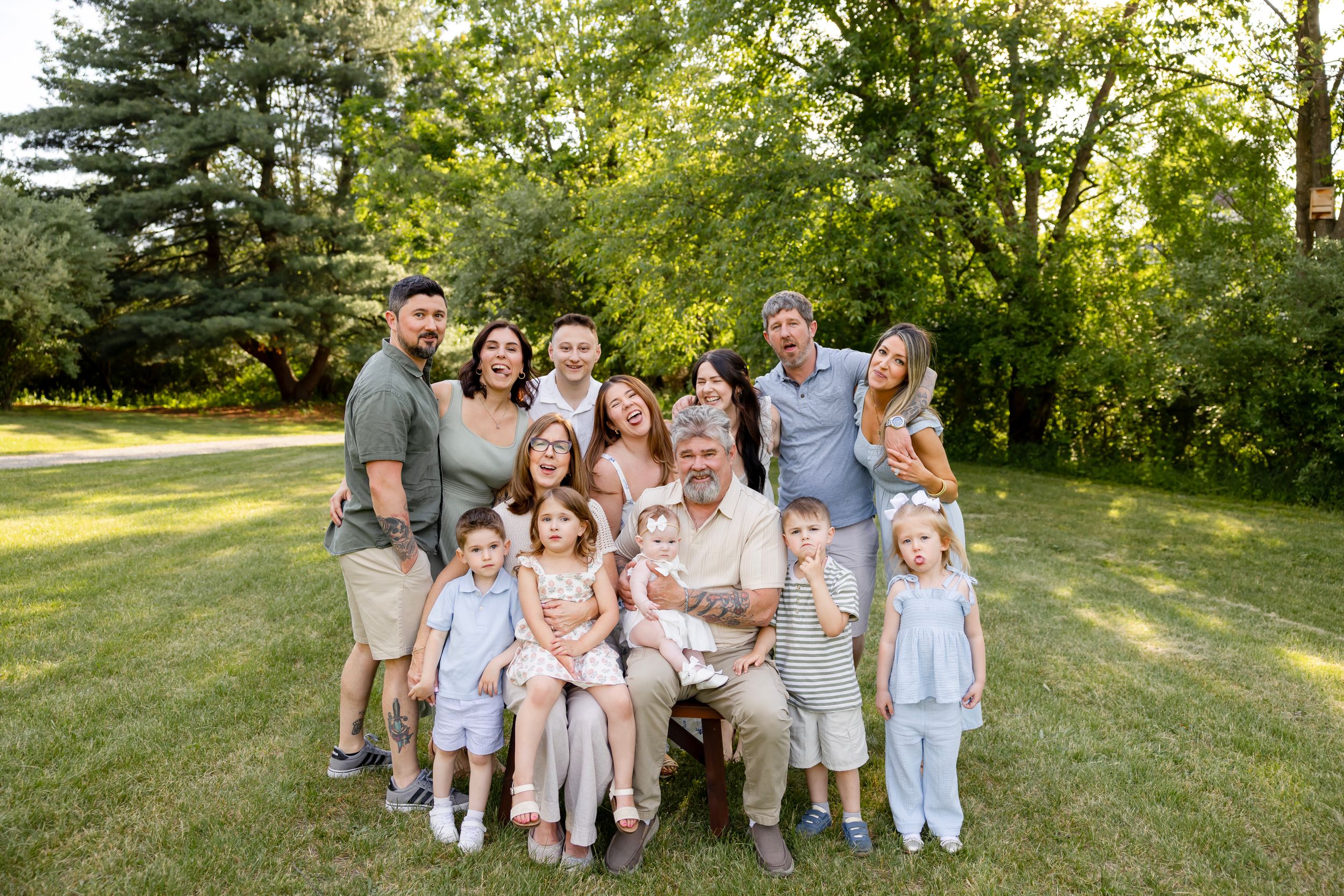 Large multigenerational family smiling together outdoors on green grass surrounded by lush trees during a sunny day.