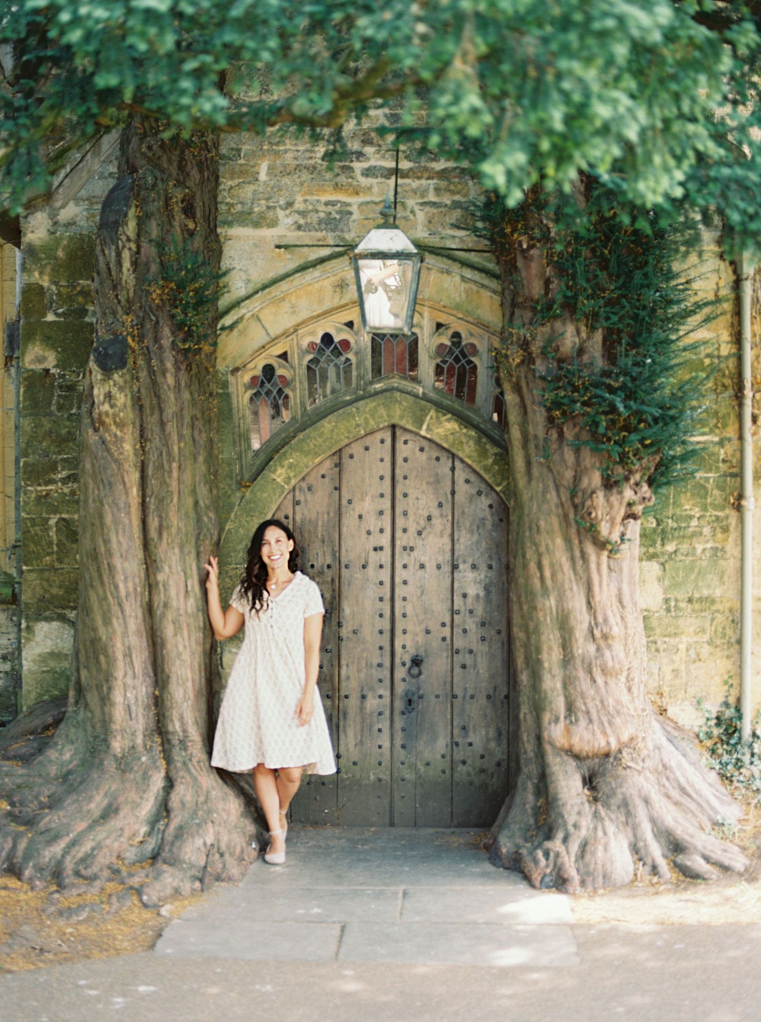 Woman in white dress poses by ancient arched wooden door flanked by large trees at a historic stone building.