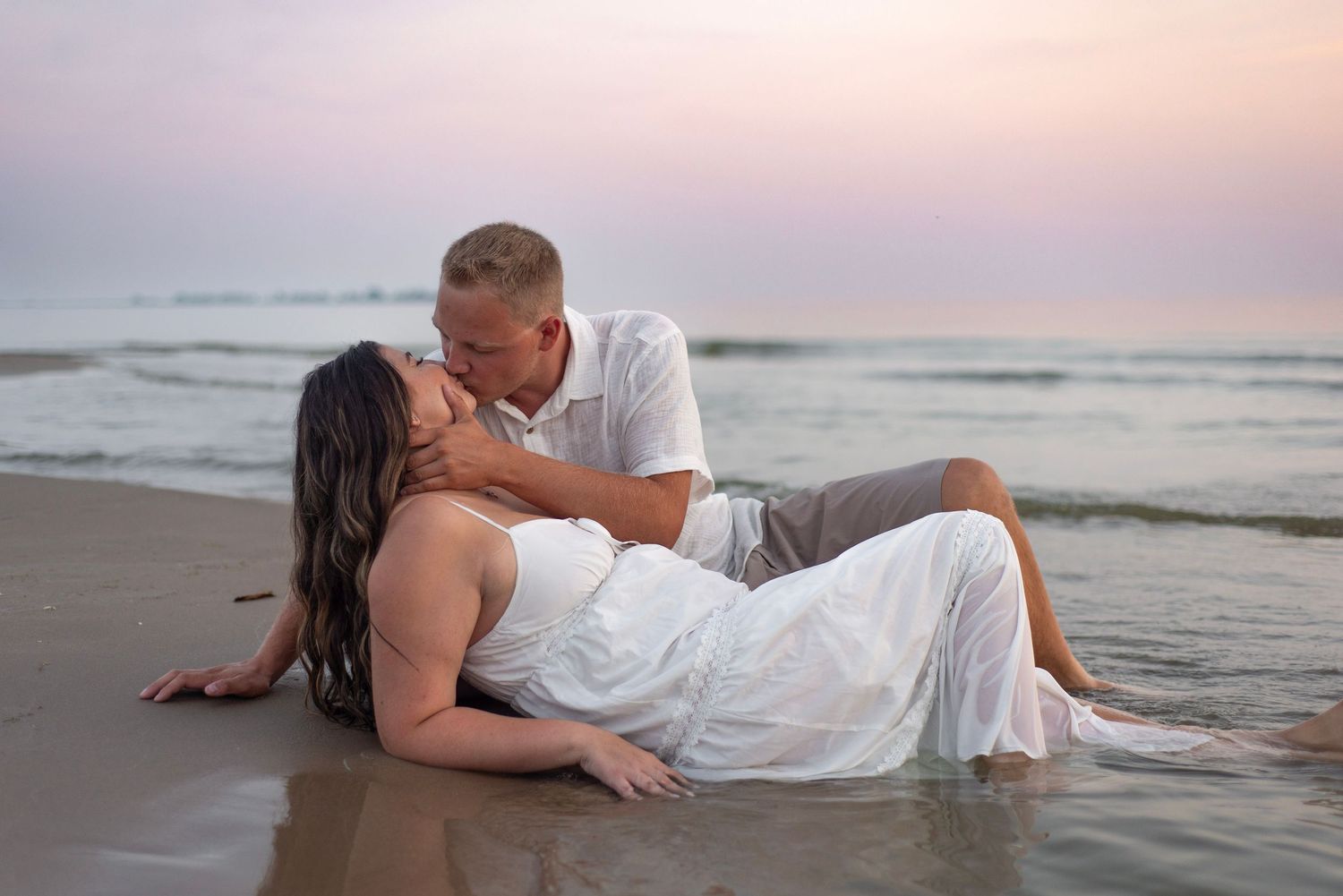 Couple sharing a romantic kiss on a sandy beach at sunset, woman in white dress lying in the shallow waves.