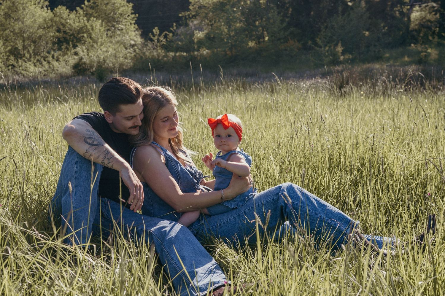 A young family sits together in a lush green field, parents smiling at their baby girl wearing a red bow.