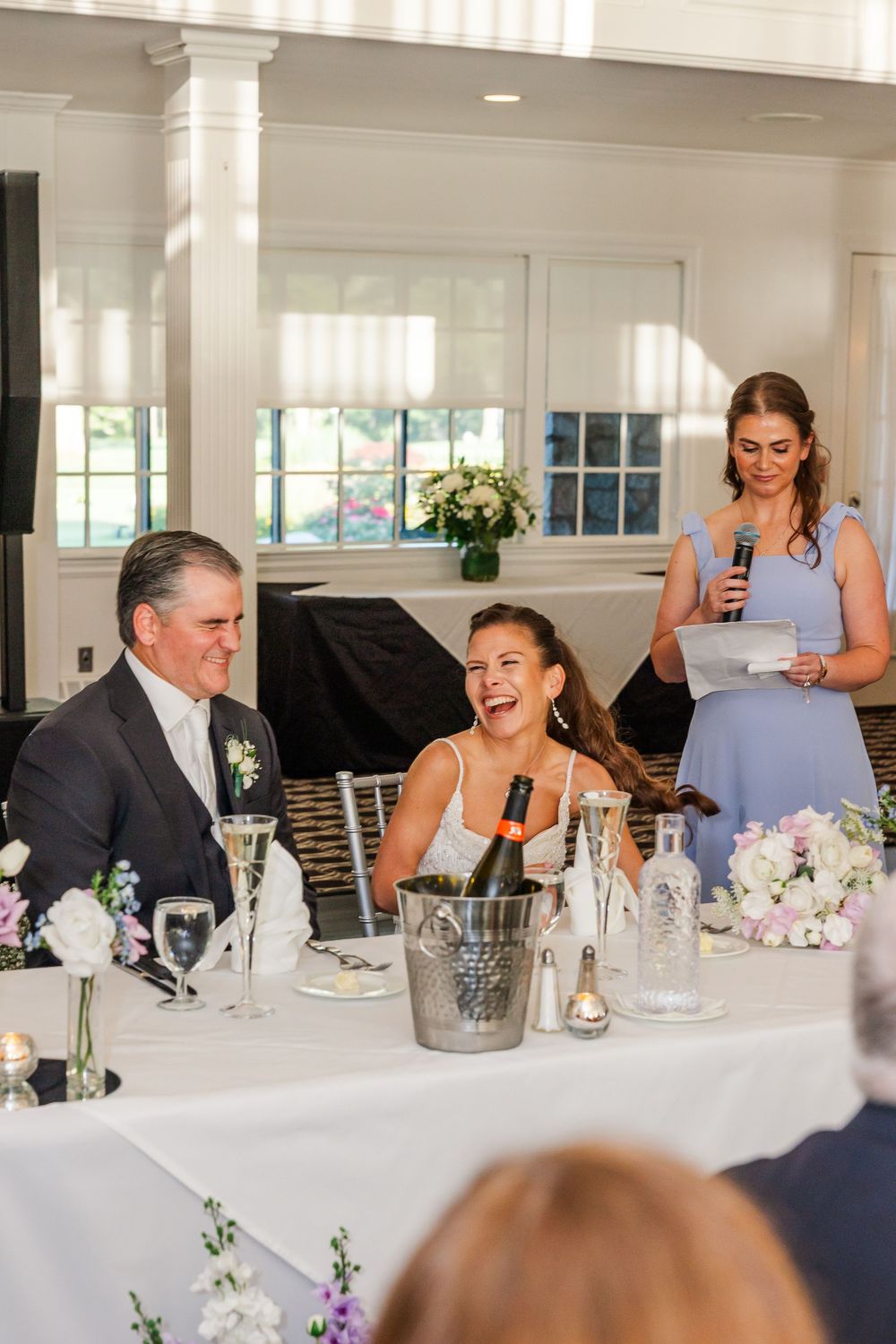 Bride laughing joyfully at wedding reception table while bridesmaid in blue gives a toast with microphone.