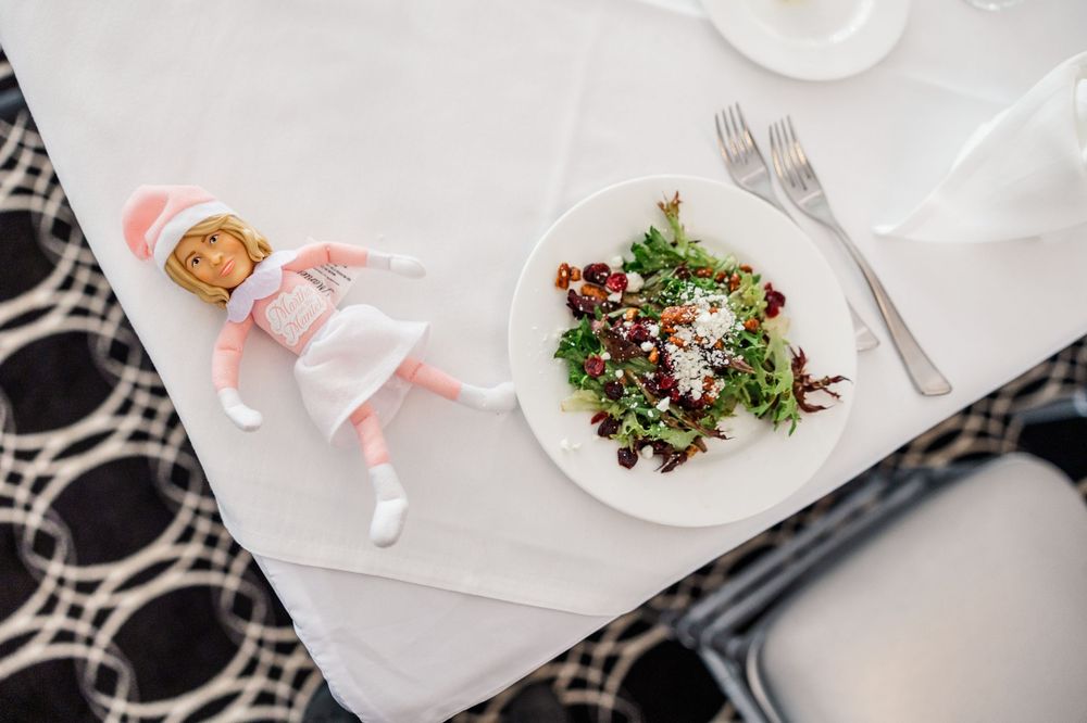 Elf on the Shelf doll lying next to a fresh salad plate with fork and knife on a white tablecloth.