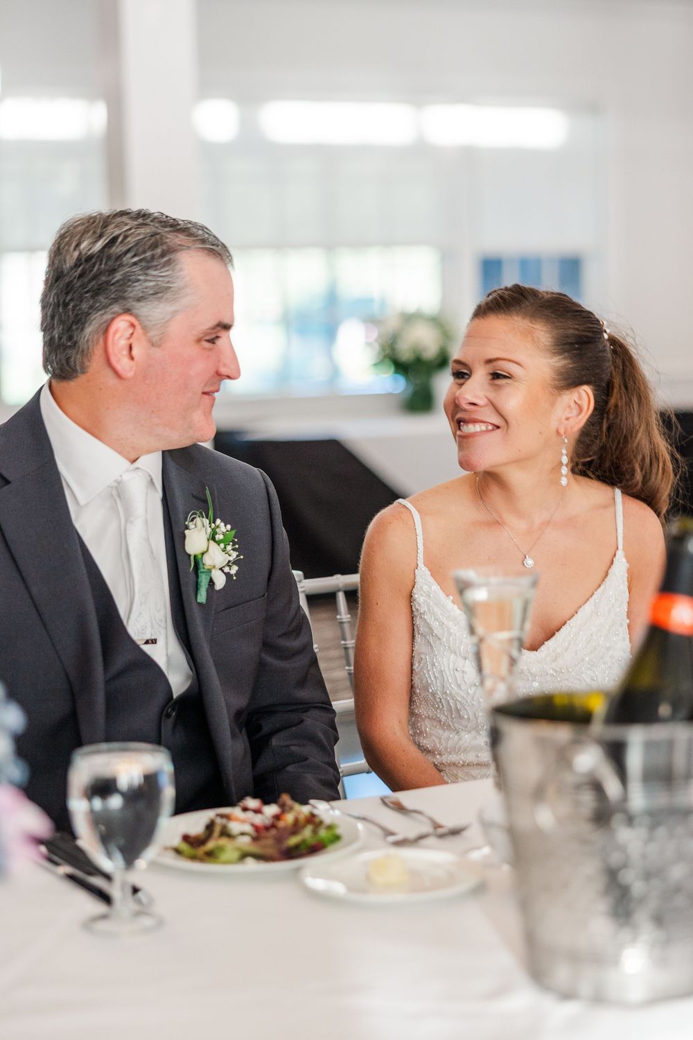 Bride and groom share a joyful moment at their wedding reception dinner table, with champagne and salads nearby.