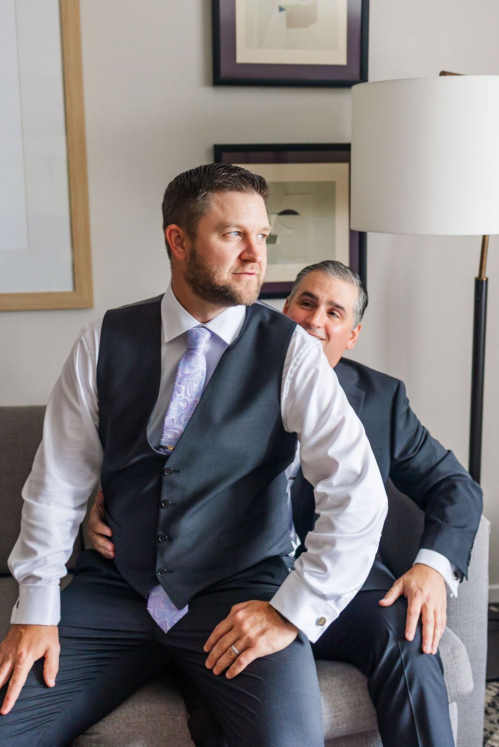 Two men in dark suits sitting together in a well-lit room, one wearing a vest and lavender tie.