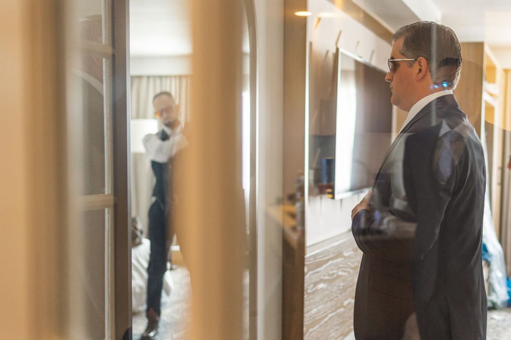 Groom in a navy suit adjusting his tie while looking at his reflection in a hotel room mirror.
