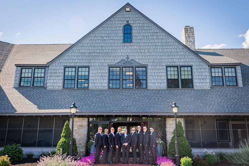 Wedding party groomsmen pose in gray suits outside a shingle-style venue with manicured gardens.