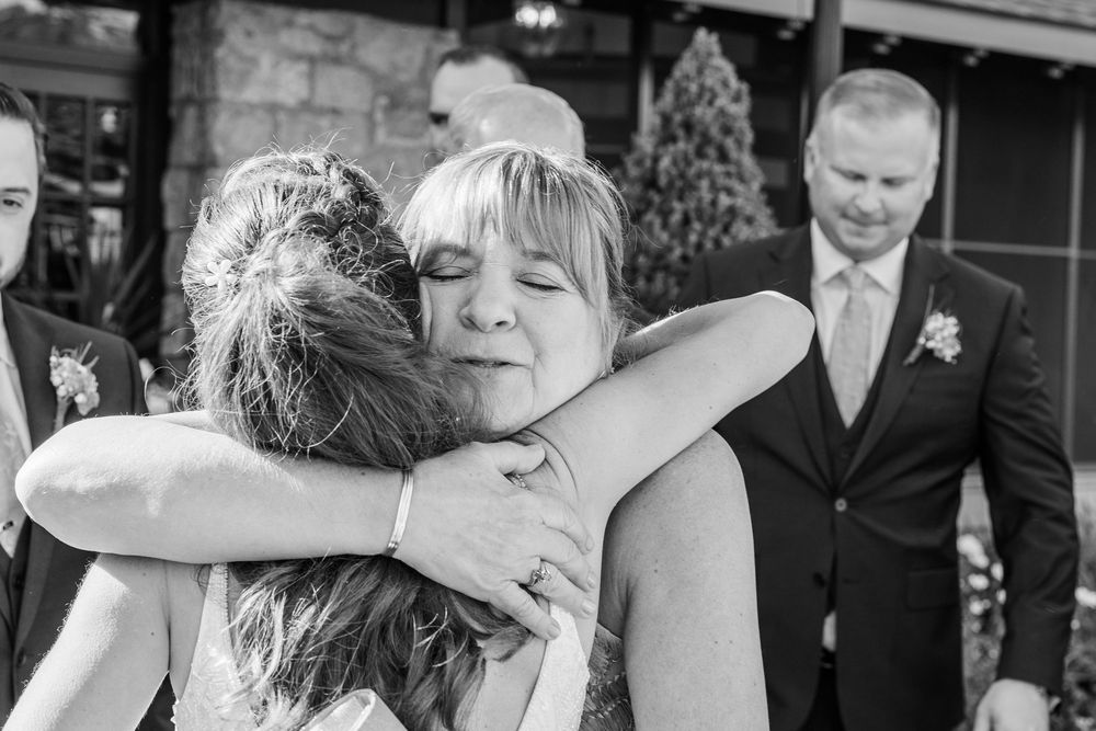 Black and white photo of two women sharing an emotional hug at a wedding, with a man in a suit visible in the background.