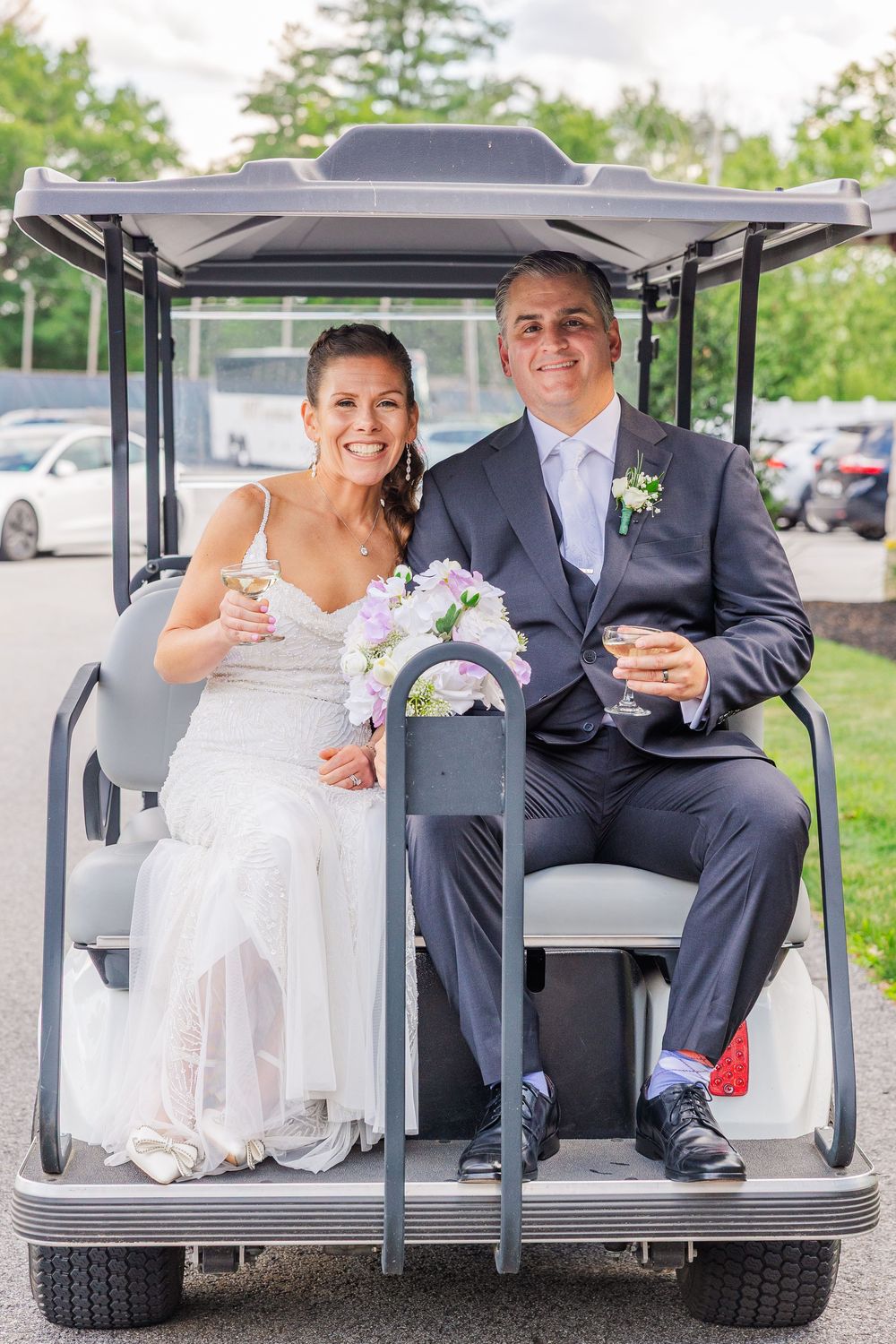Smiling bride and groom ride a golf cart holding champagne glasses at their wedding.