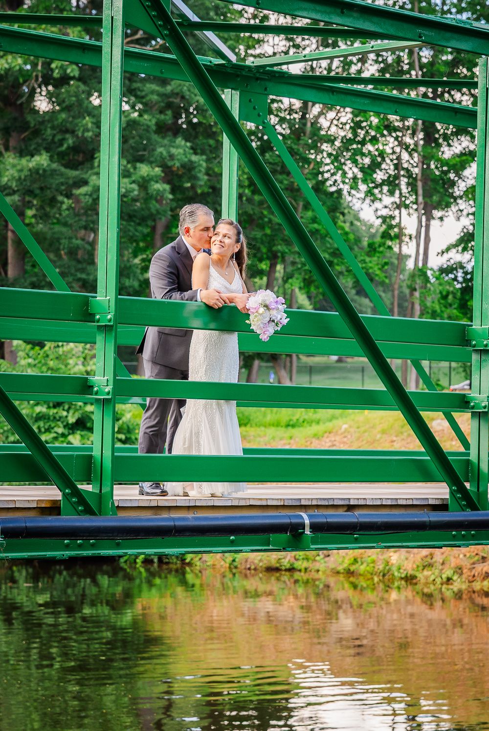 Bride and groom share a tender kiss on a green steel bridge over a pond.