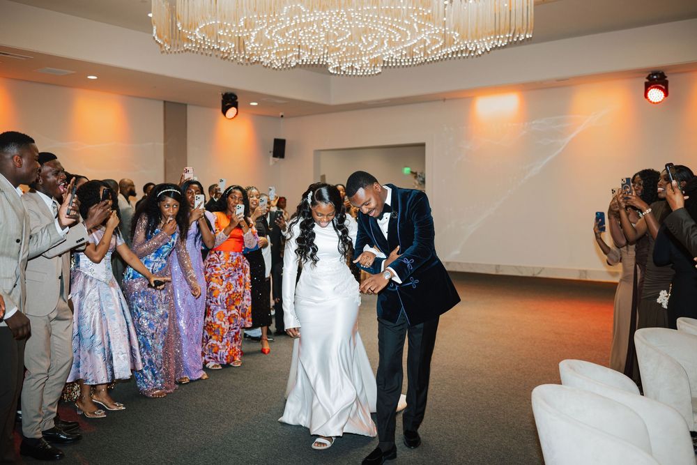 Bride and groom share first dance at elegant wedding reception with guests cheering and crystal chandelier overhead.