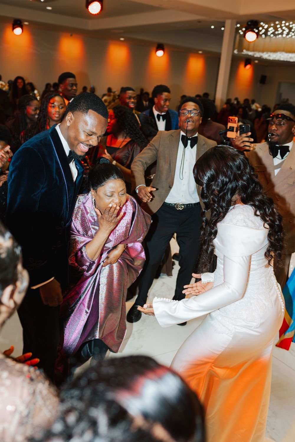 Emotional wedding moment as bride kneels before surprised guest in white gown at elegant reception venue.
