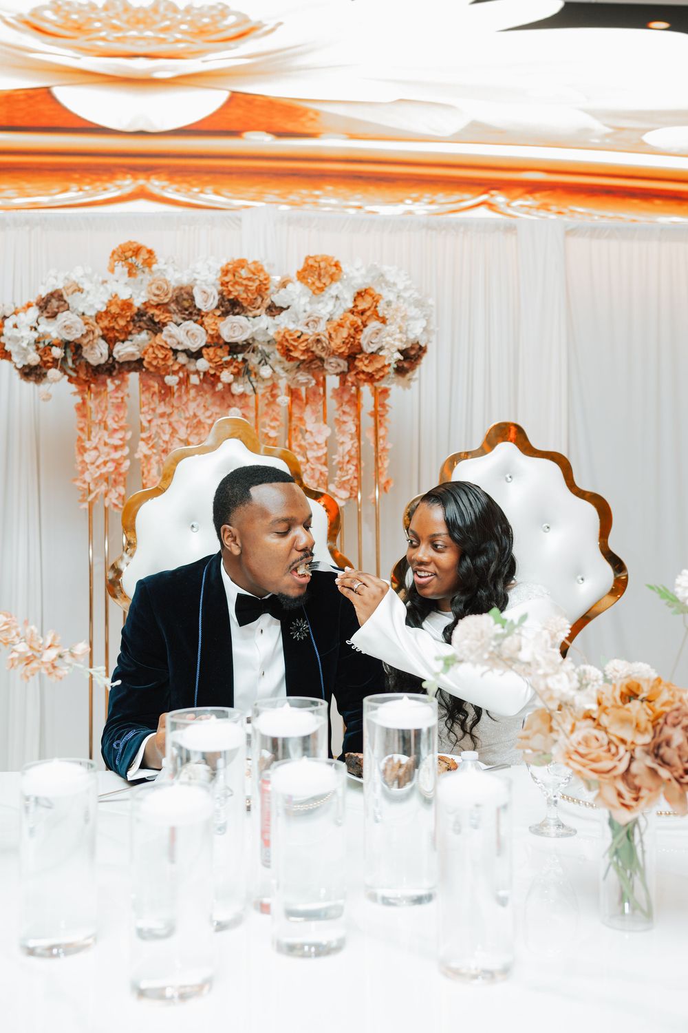Bride feeds groom at elegant wedding reception with gold throne chairs and orange floral backdrop decorations.