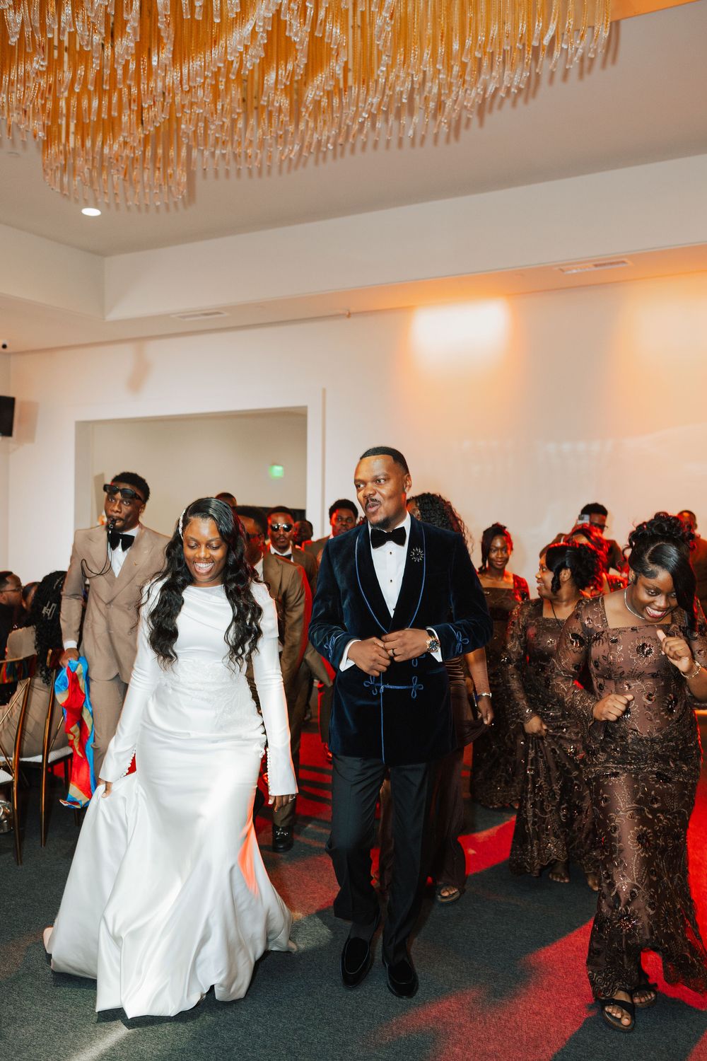 Smiling bride in white gown walks with groom in navy velvet tuxedo during wedding reception entrance.