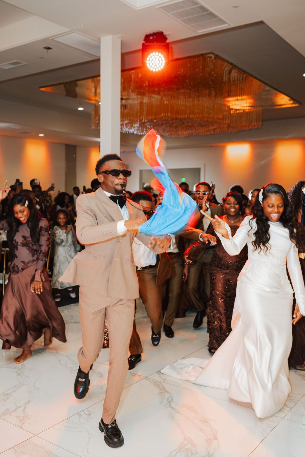Groom in tan suit and sunglasses dances joyfully with bride in white gown at lively wedding reception.