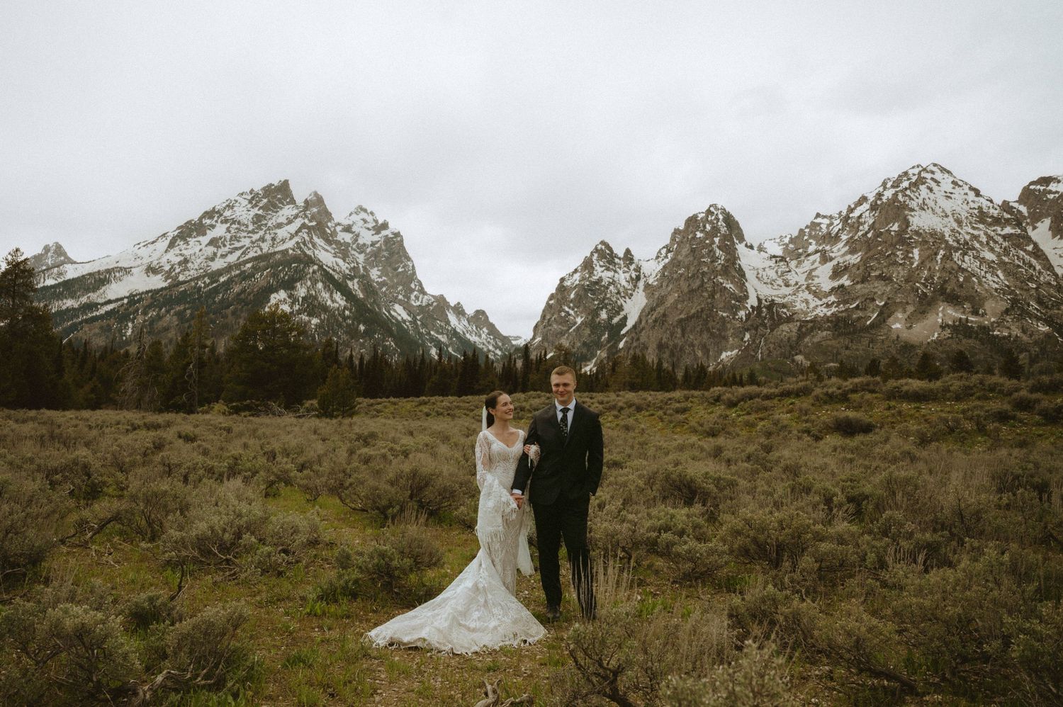 Bride and groom stand in mountain meadow with snow-capped Grand Teton peaks behind them on cloudy day.