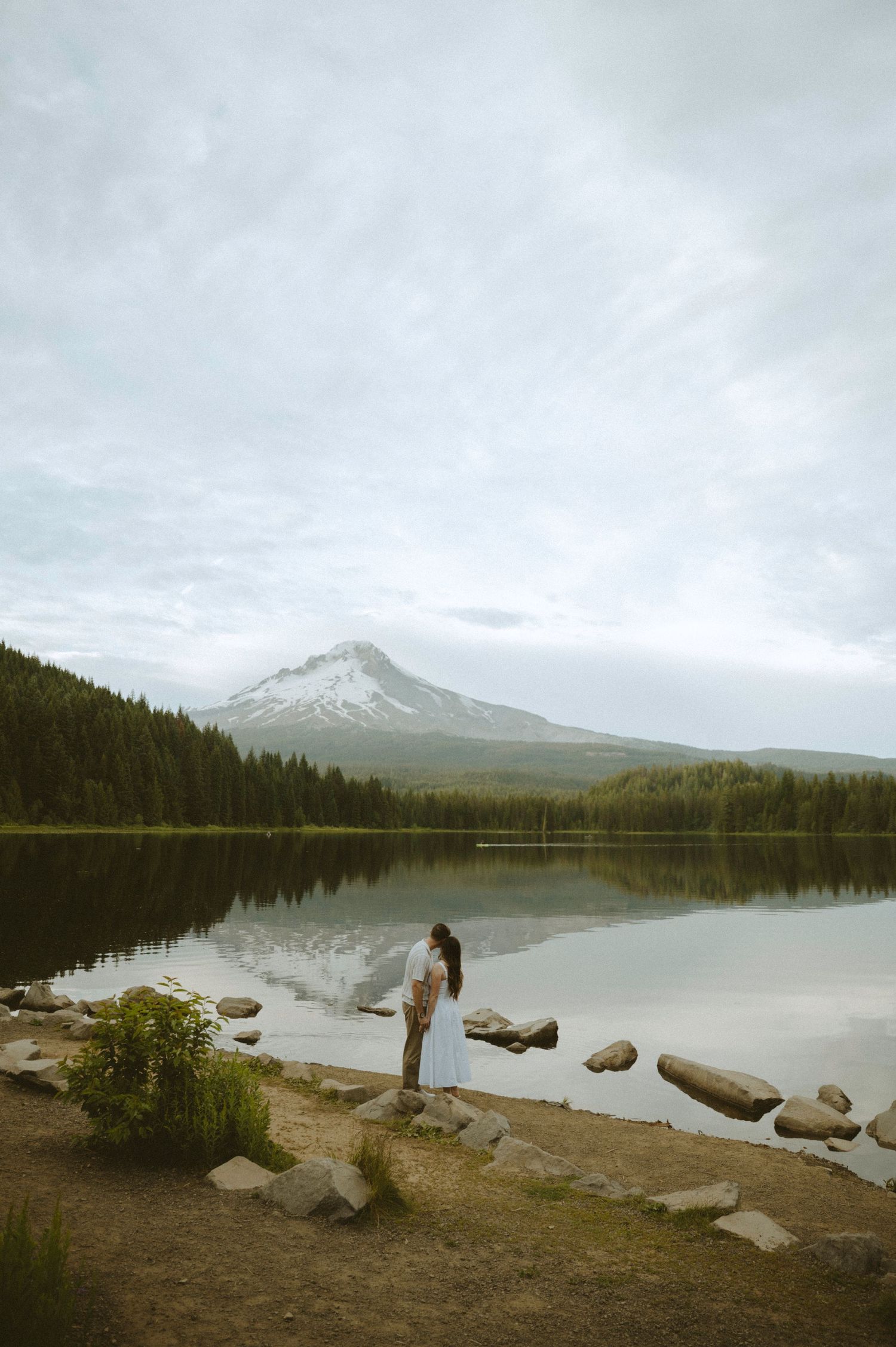 Couple standing by a serene mountain lake with Mt. Hood reflected in calm waters, surrounded by evergreen forest.