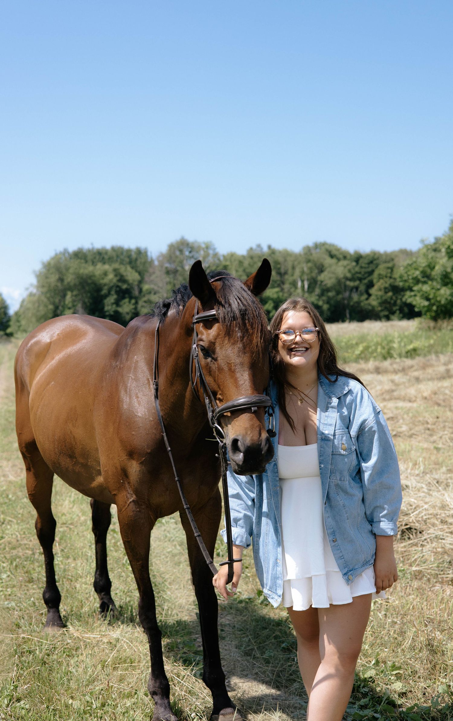 Smiling girl in white dress and denim jacket posing with a bay horse in a sunny field.