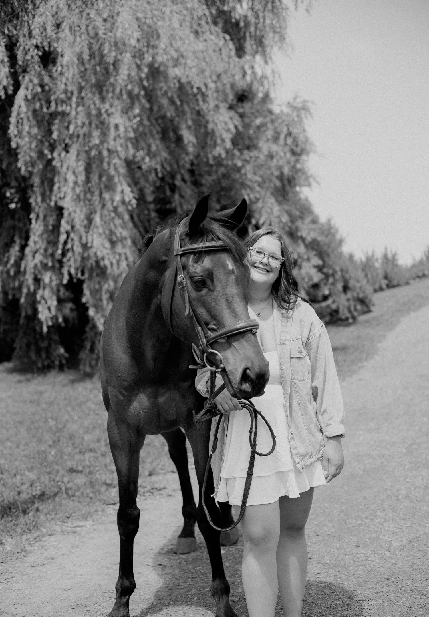 Smiling girl with glasses standing beside a bay horse under a willow tree in black and white.