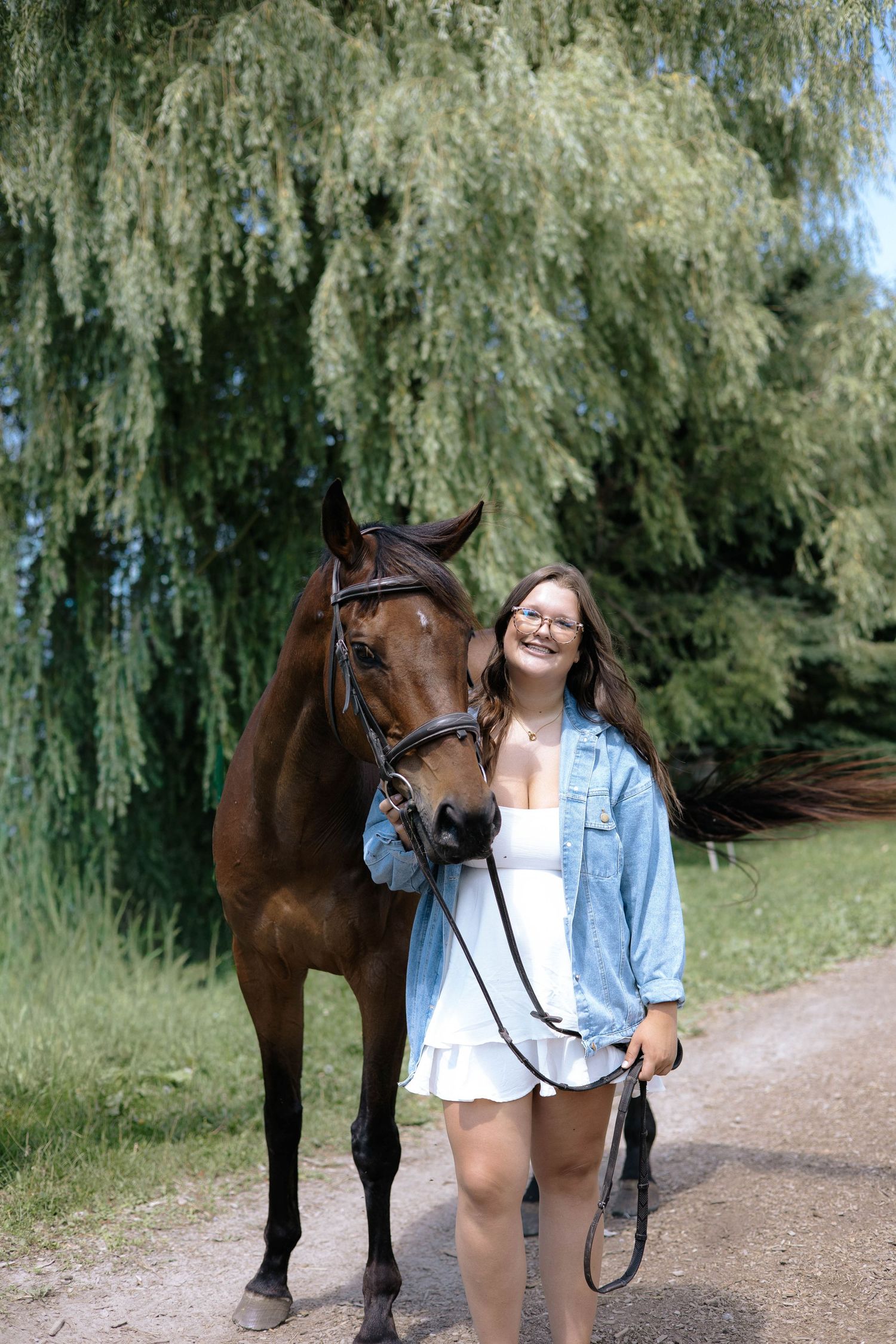 Young woman in denim jacket walking beside a bay horse on a gravel path by a willow tree.