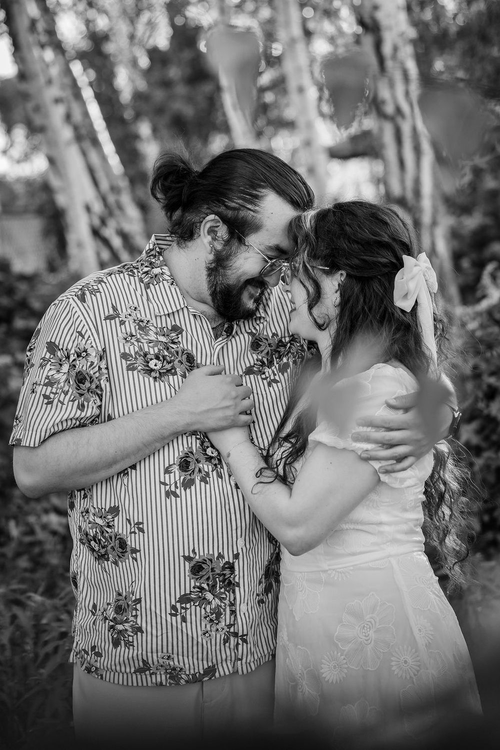 Black and white photo of a bearded groom in floral shirt touching foreheads with bride in white dress outdoors.