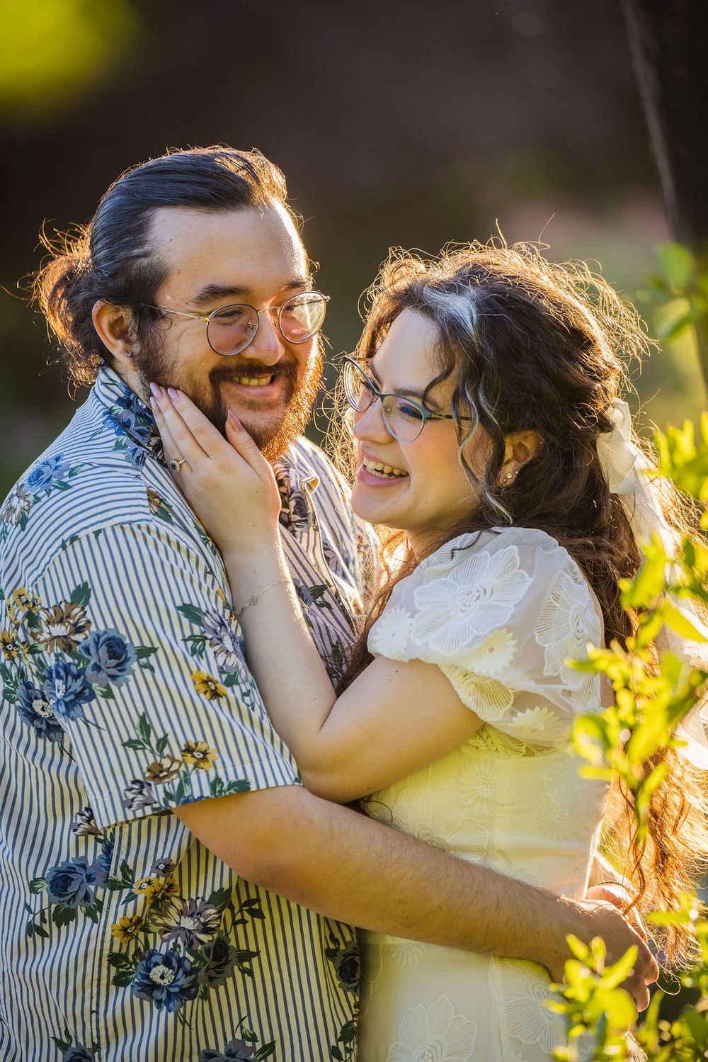 Couple laughing and embracing outdoors in golden sunlight, woman in yellow dress touching mans bearded face.