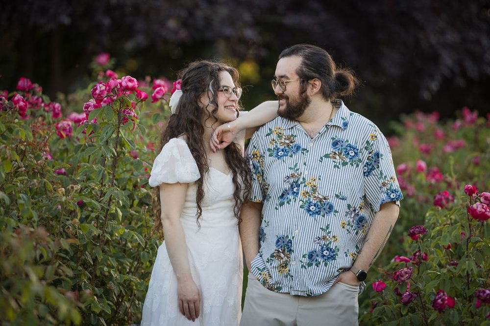 Happy couple standing together among pink roses in a garden, woman in white dress smiling up at bearded man.