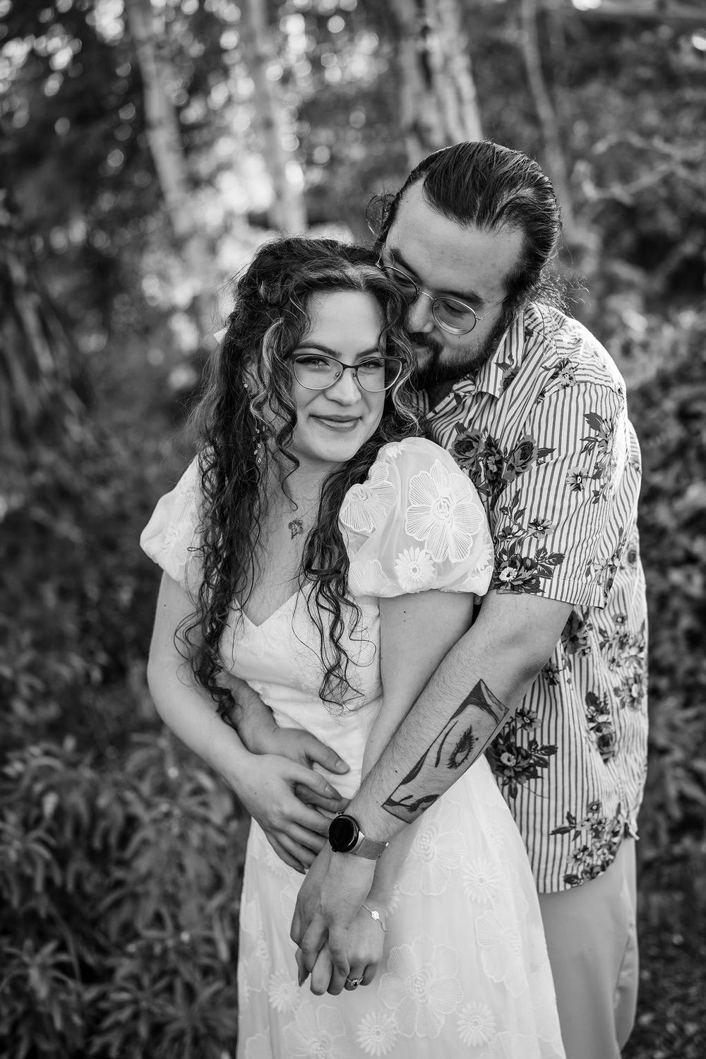 Black and white photo of a couple embracing outdoors, man kissing womans cheek, she smiles wearing glasses and white dress.