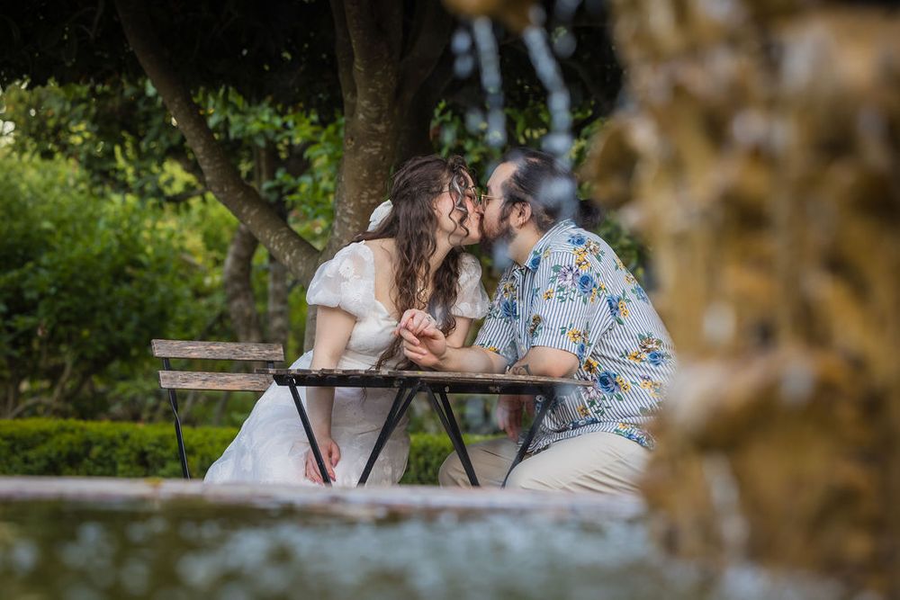 Couple sharing a romantic kiss at a garden bench near a fountain, surrounded by lush green trees.